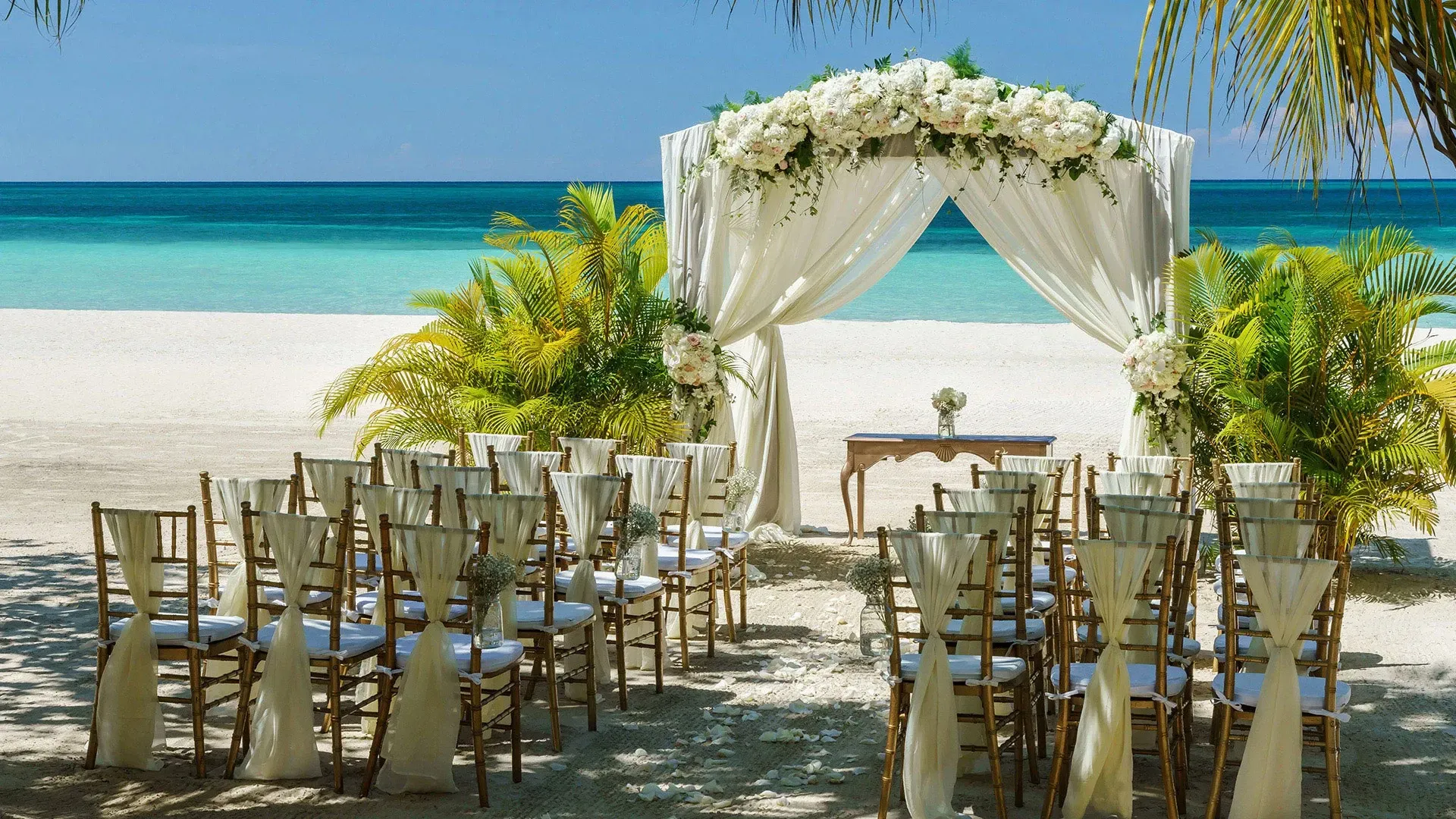 Elegant beachfront wedding ceremony setup with white floral arch and wooden chairs.
