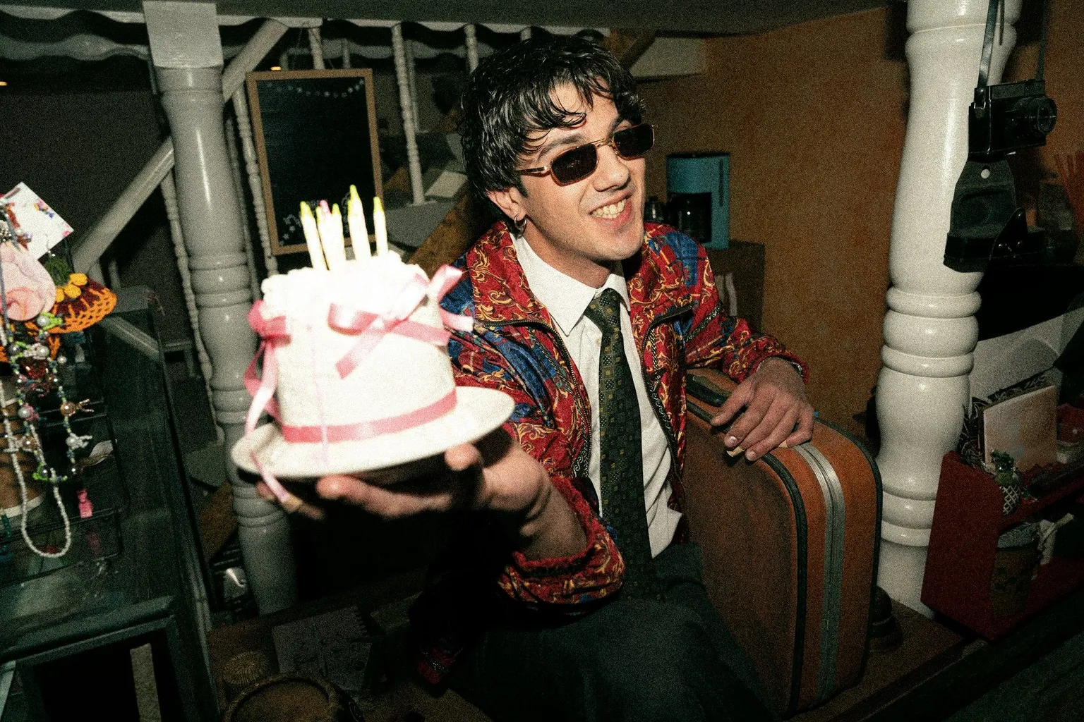 Young man wearing sunglasses and holding out a small birthday cake with lit candles.