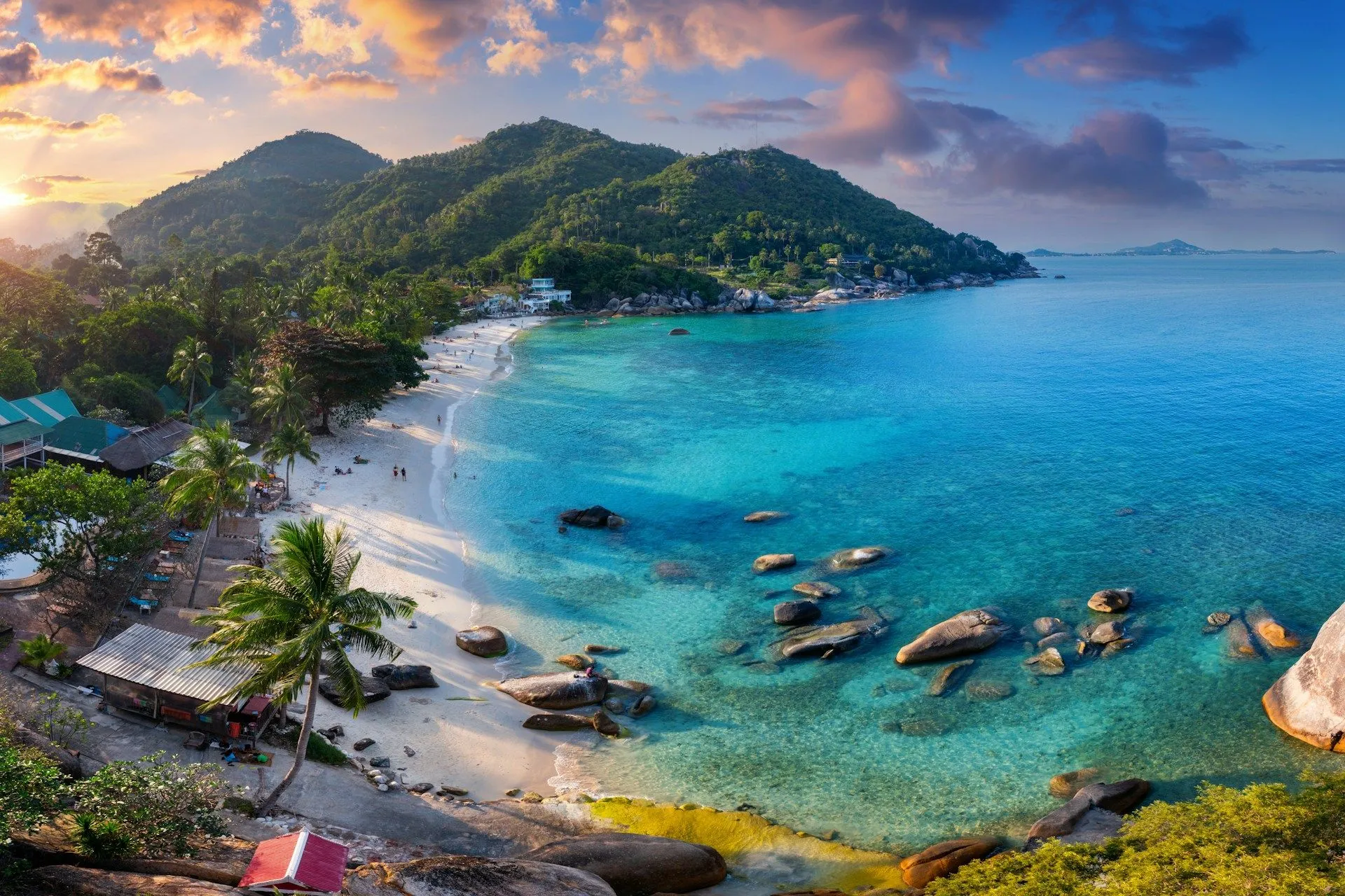 Aerial view of a tropical beach with turquoise water, rocky shoreline, and lush green hills.