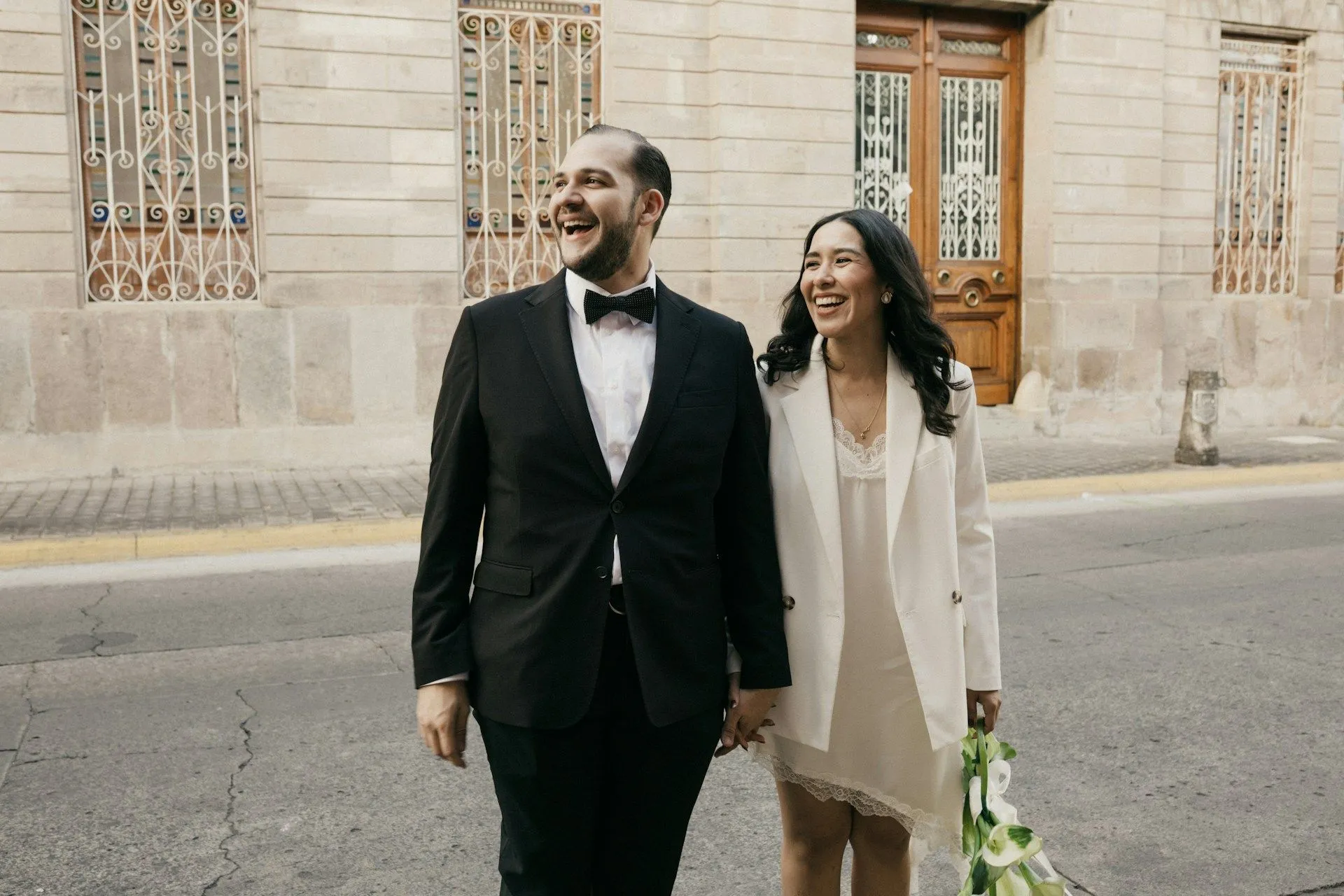 Smiling couple in formal attire walking hand in hand along a city street.